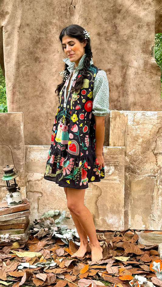 Side view of a woman wearing a black short dress with a vibrant illustrated print, against a natural rustic backdrop