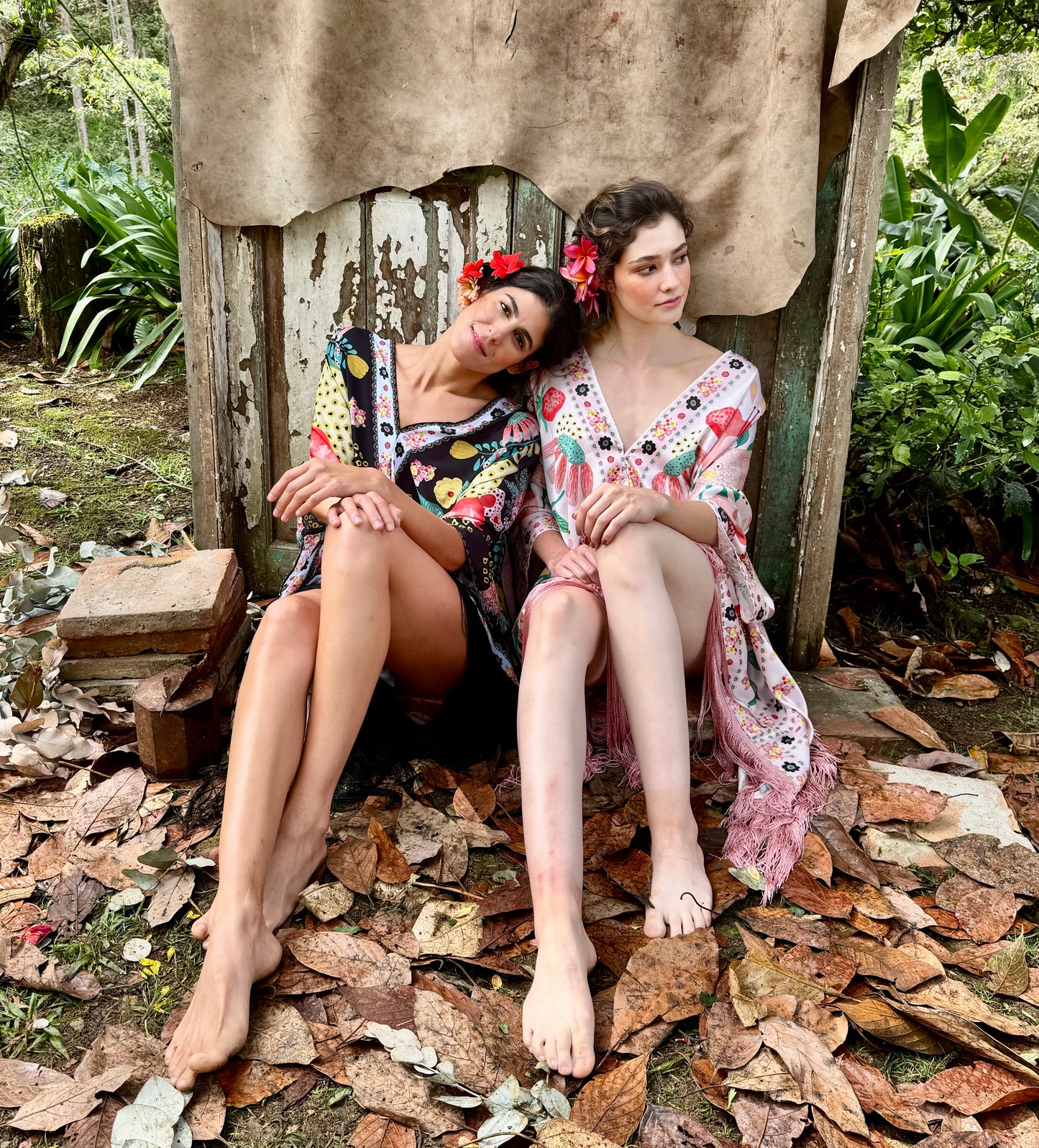 Two women seated in nature wearing a printed a black and pink TelaCuento kimono, showcasing its draped chiffon fabric and long fringe