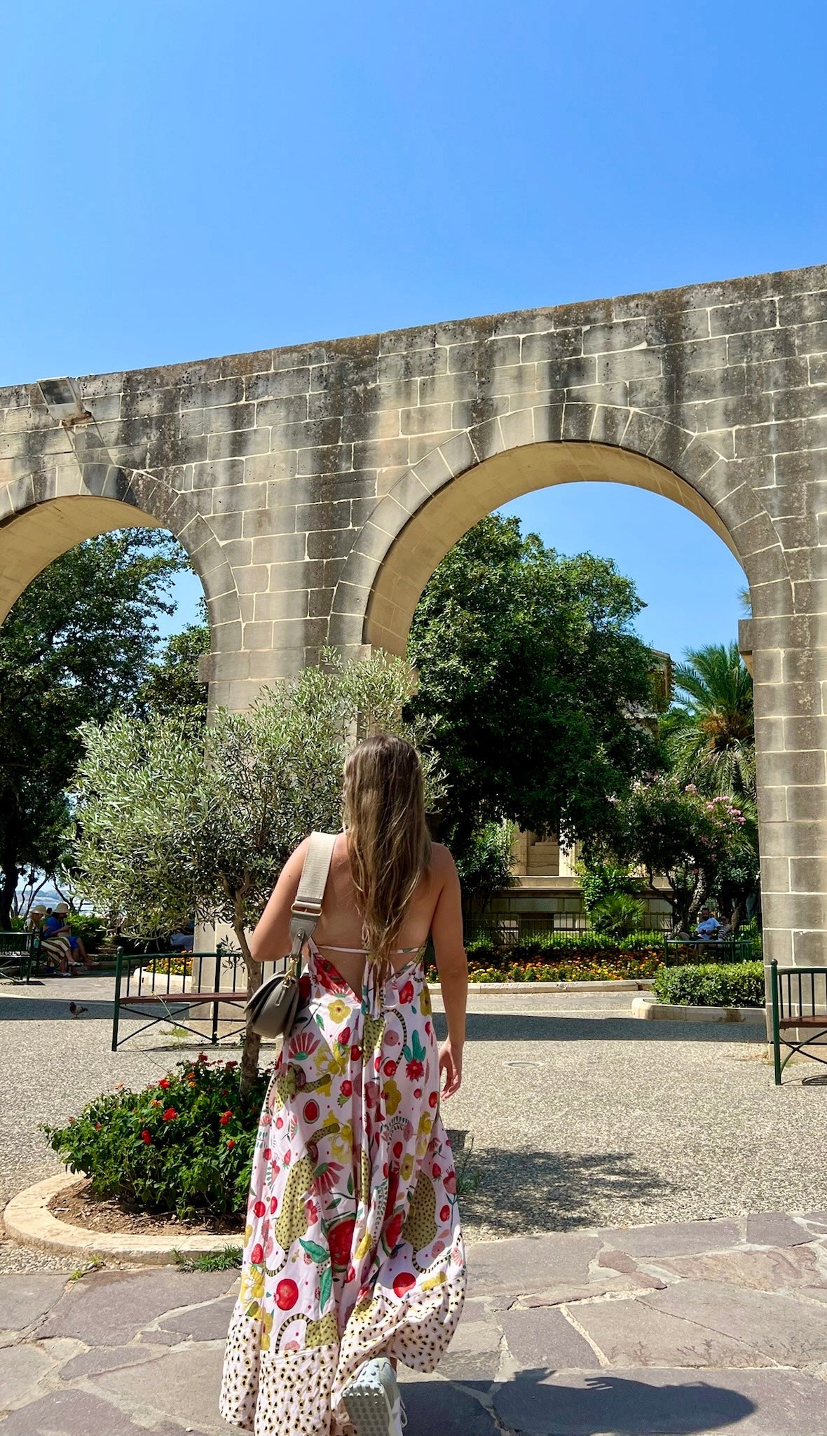 Woman in a floral dress standing under a stone archway with a clear blue sky.