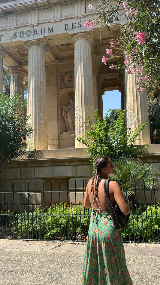Back view of a woman wearing the green botanical halter-neck dress, standing in front of stone columns and pink flowering plants.