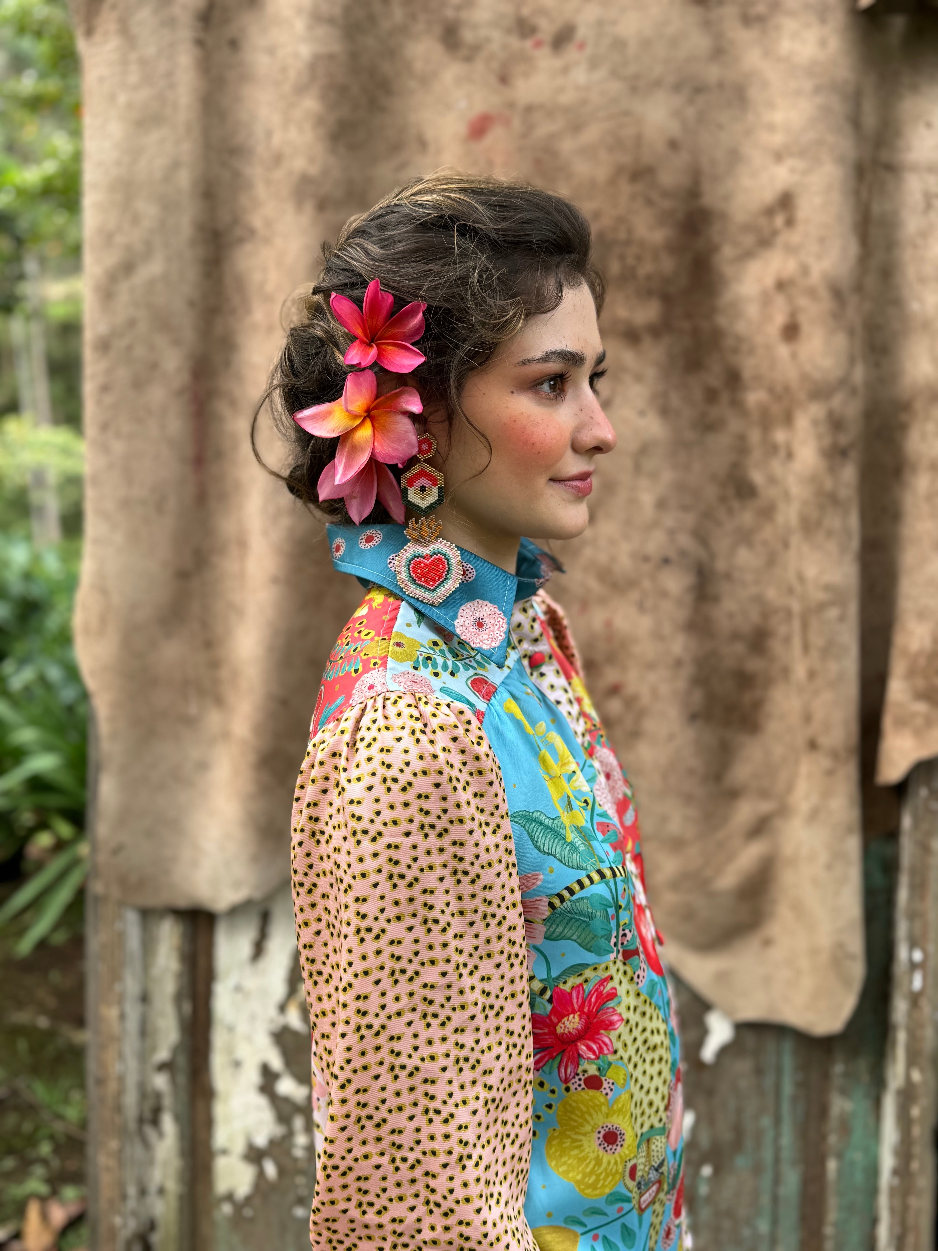 Woman standing outdoors wearing a colourful printed long-sleeve chiffon shirt, styled with floral accessories in her hair, against a textured natural background.