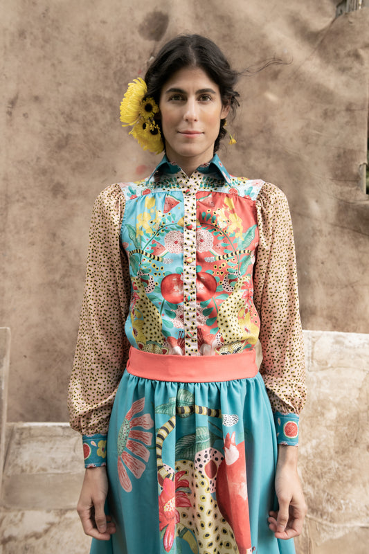 Front view of a woman wearing a vibrant hand-illustrated long-sleeve chiffon shirt paired with a matching skirt, standing against a rustic backdrop.