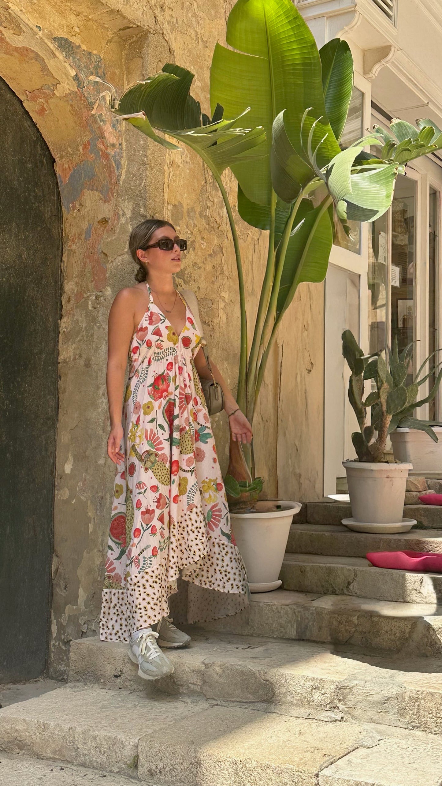 Woman in a floral dress standing on stone steps with plants and architectural elements in the background